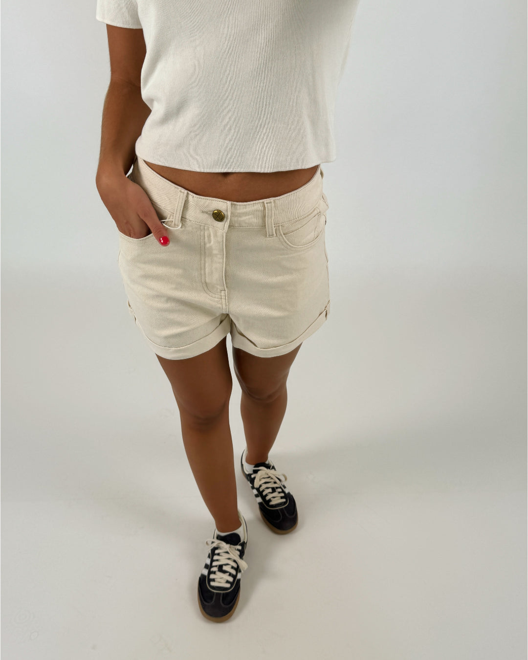 Person iført lyse shorts og hvid t-shirt står med hånden i lommen og iført mørke sneakers. Baggrunden er neutral og lys, hvilket fremhæver tøjet.