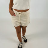 Person iført lyse shorts og hvid t-shirt står med hånden i lommen og iført mørke sneakers. Baggrunden er neutral og lys, hvilket fremhæver tøjet.