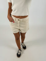 Person iført lyse shorts og hvid t-shirt står med hånden i lommen og iført mørke sneakers. Baggrunden er neutral og lys, hvilket fremhæver tøjet.