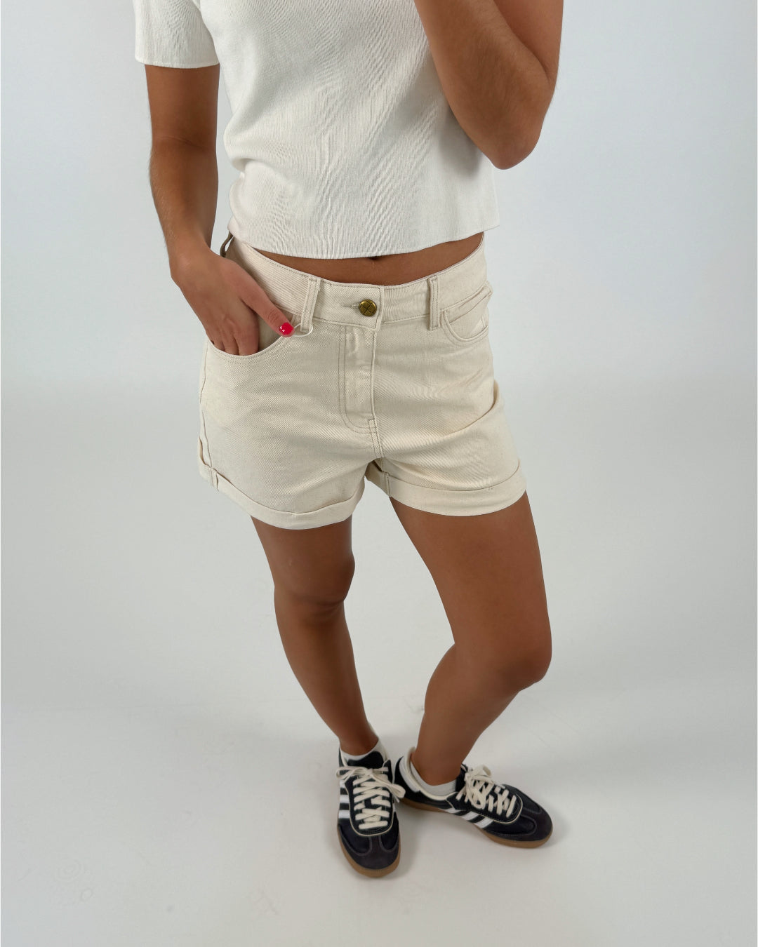 En person iført en hvid kortærmet trøje og beige shorts står med den ene hånd i lommen. Personen har sorte sneakers på. Baggrunden er neutral og hvid.