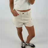 En person iført en hvid kortærmet trøje og beige shorts står med den ene hånd i lommen. Personen har sorte sneakers på. Baggrunden er neutral og hvid.