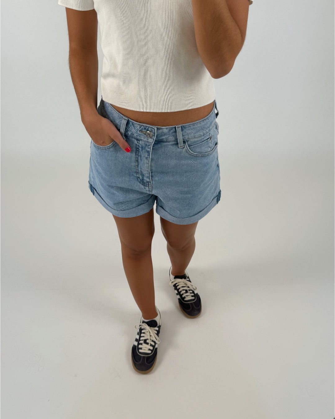 En person iført lyseblå denimshorts og en hvid t-shirt står med hånden i lommen. De bærer mørke sneakers med hvide snørebånd. Baggrunden er lys og neutral.