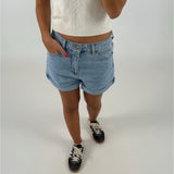 En person iført lyseblå denimshorts og en hvid t-shirt står med hånden i lommen. De bærer mørke sneakers med hvide snørebånd. Baggrunden er lys og neutral.