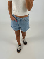En person iført lyseblå denimshorts og en hvid t-shirt står med hånden i lommen. De bærer mørke sneakers med hvide snørebånd. Baggrunden er lys og neutral.