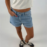 En person iført lyseblå denimshorts og hvide sneakers står med hænderne i lommerne på en lys, ensfarvet baggrund.