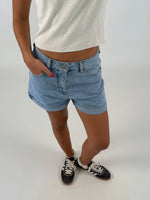 En person iført lyseblå denimshorts og hvide sneakers står med hænderne i lommerne på en lys, ensfarvet baggrund.