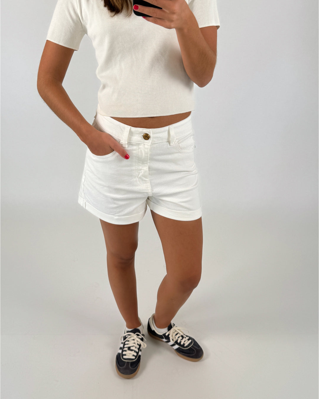 En person i en hvid t-shirt og shorts tager et billede, mens de står på et lyst gulv, iført sorte sneakers med hvide snørebånd.