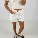 En person i en hvid t-shirt og shorts tager et billede, mens de står på et lyst gulv, iført sorte sneakers med hvide snørebånd.
