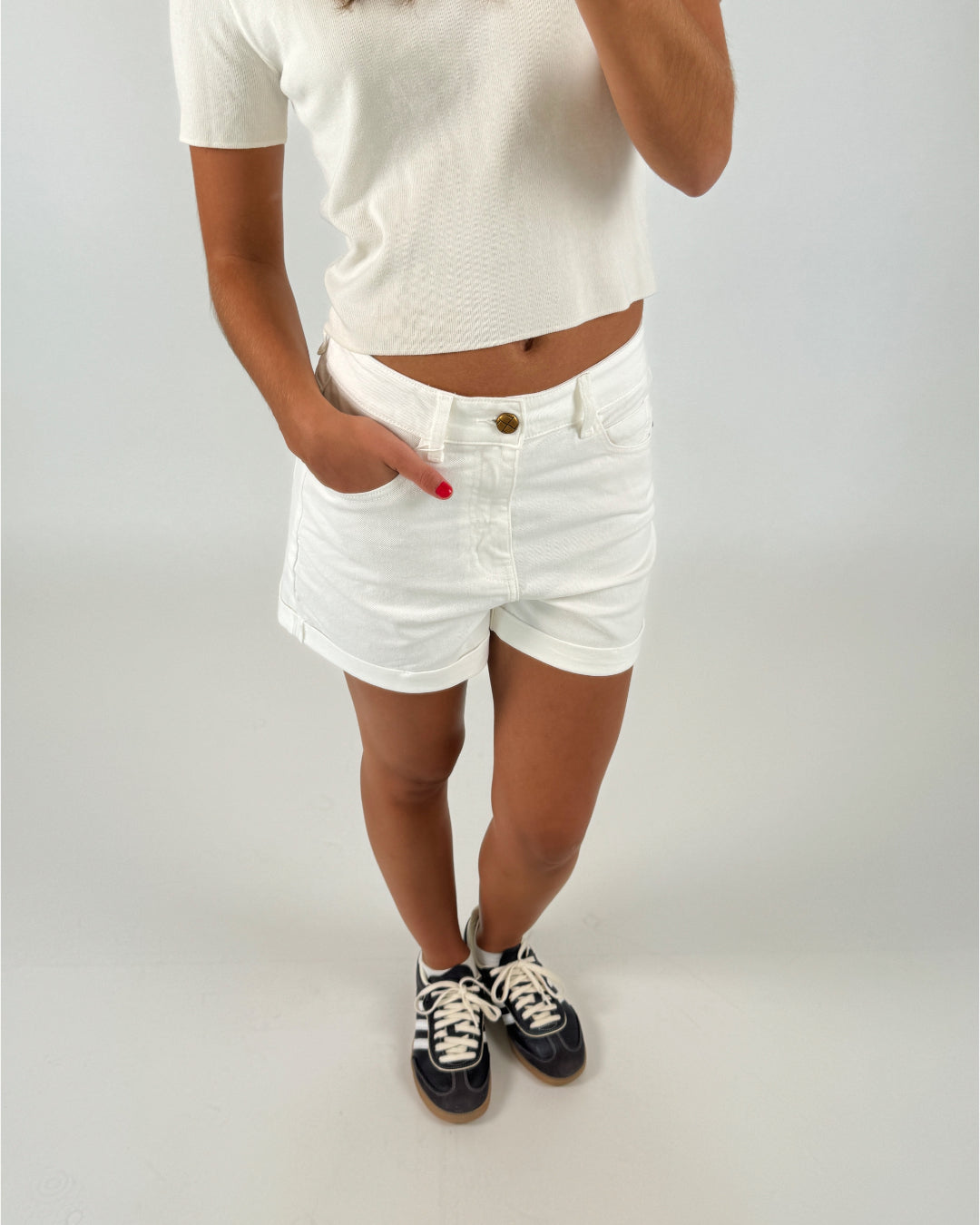 En person iført en hvid t-shirt og shorts står med hånden i lommen, iført sorte sneakers med lyse snørebånd på en lys baggrund.