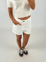 En person iført en hvid t-shirt og shorts står med hånden i lommen, iført sorte sneakers med lyse snørebånd på en lys baggrund.