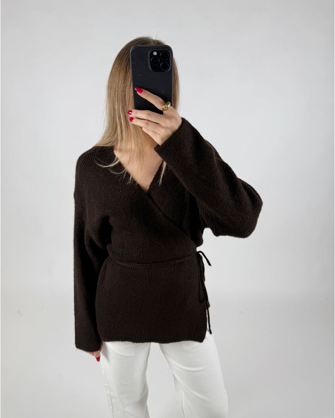 Person i mørk sweater holder en smartphone foran ansigtet; baggrunden er en ensfarvet væg. Hun bærer hvide bukser og har rødlakerede negle.