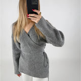 En person iført en grå sweater tager et spejlbillede med en sort mobiltelefon; personen har langt, lyst hår og rødmalede negle, stående i et enkelt, hvidt rum.