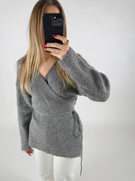 En person iført en grå sweater tager et spejlbillede med en sort mobiltelefon; personen har langt, lyst hår og rødmalede negle, stående i et enkelt, hvidt rum.