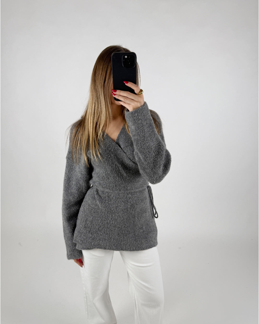 En person tager et billede med en telefon iført en grå trøje og hvide bukser, stående i et minimalistisk, lyst rum.