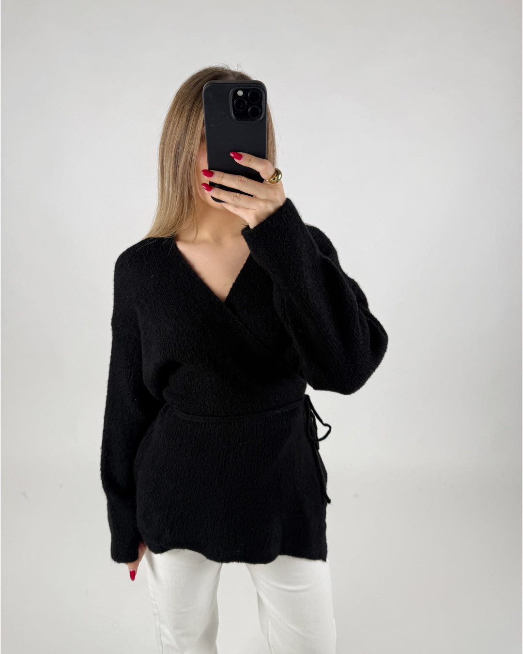 Person tager et billede med en smartphone, iført en sort sweater og hvide bukser. Baggrunden er enkel og hvid, hvilket fremhæver tøjet og personen tydeligt.