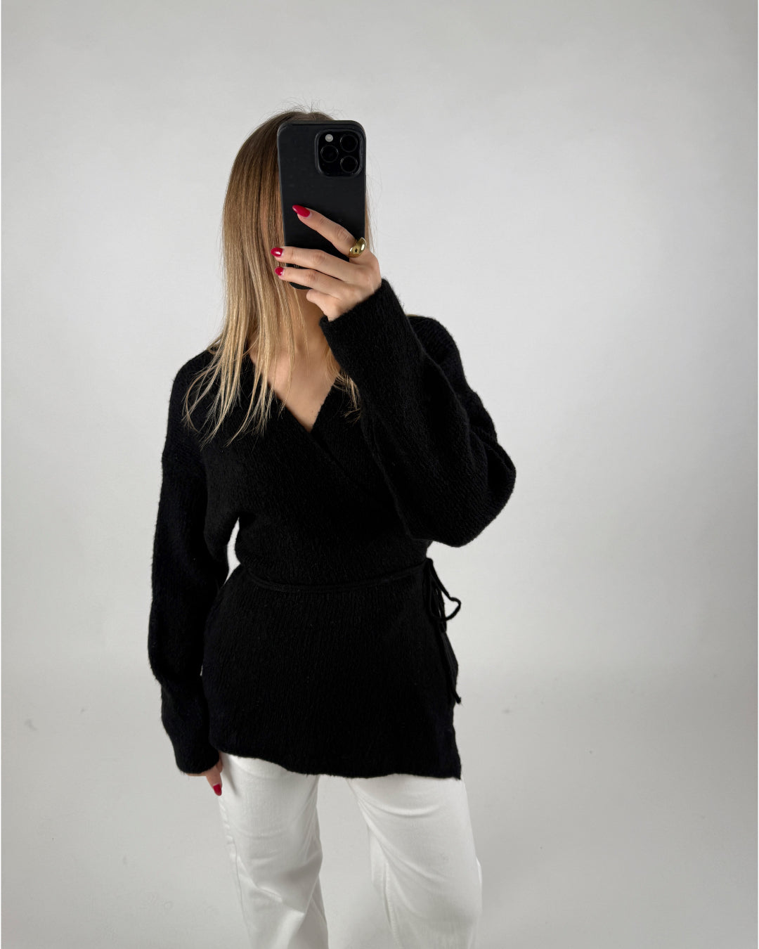 En person tager et spejlbillede med en telefon, iført en sort trøje og hvide bukser, mod en ensfarvet baggrund.