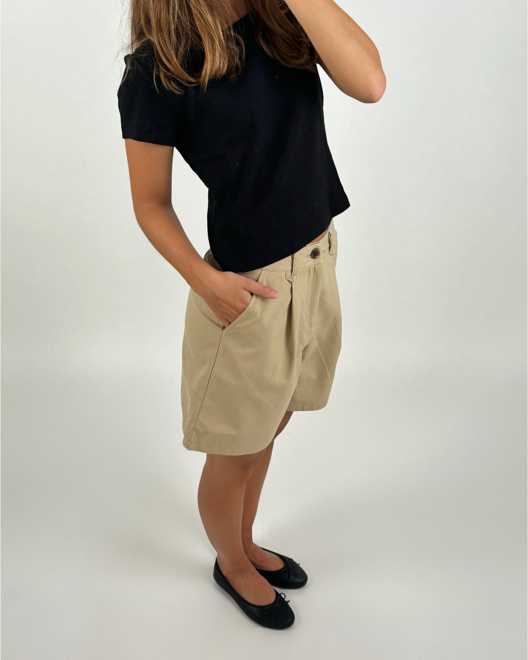 Person iført sort t-shirt og beige shorts står med hånden i lommen i et lyst rum, ansigtet delvis skjult af hænder.