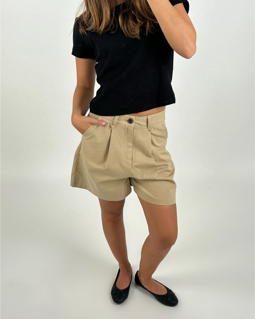 Person iført sort t-shirt og beige shorts har en hånd i lommen, står på lys baggrund.