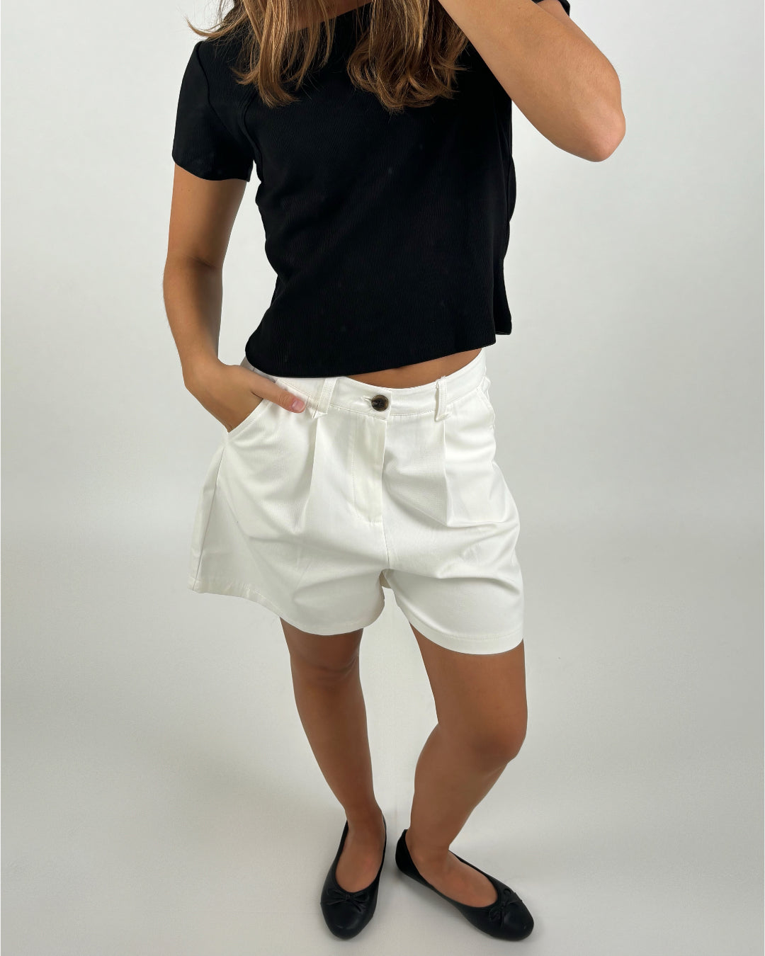 En person i en sort t-shirt og hvide shorts står med den ene hånd i lommen i et neutralt, lyst miljø. Personen bærer sorte sko.