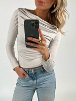 En person med hvid bluse, blå jeans og smykker holder en sort smartphone, mens vedkommende tager et billede mod en neutral baggrund.