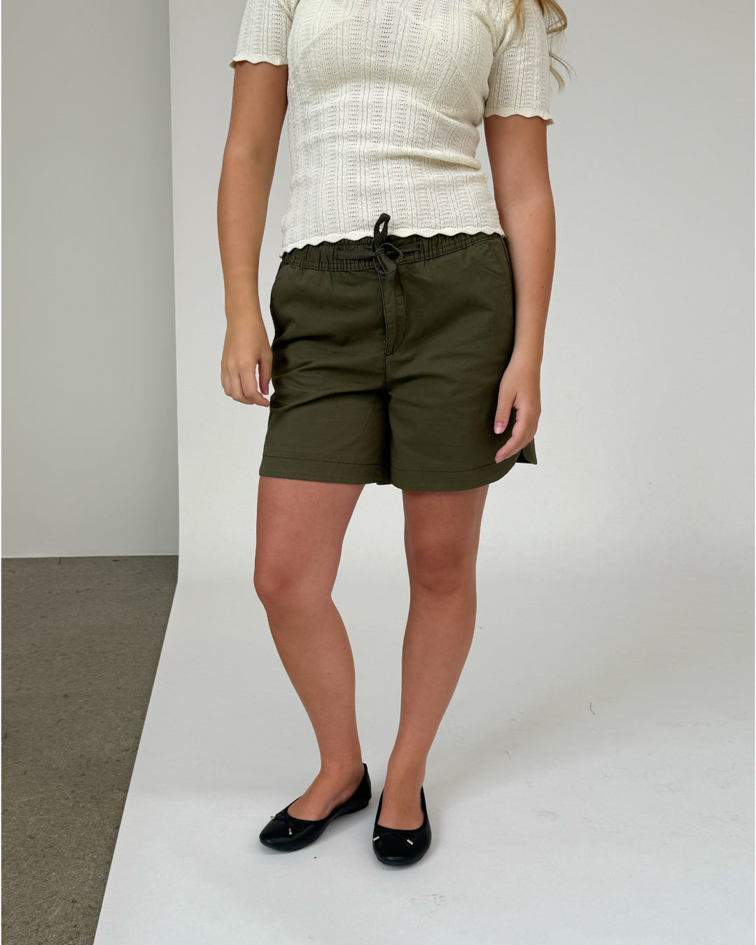 Person står iført hvid strikket bluse og grønne shorts med bindebånd, på en grå-hvid baggrund, iført sorte flade sko.