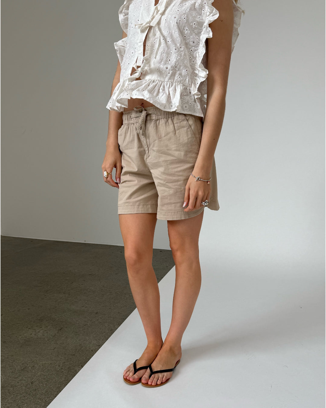 Person iført beige shorts og hvid bluse med flæser, stående på krydsfelt mellem gråt og hvidt underlag, indendørs fotograferet.