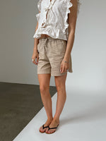 Person iført beige shorts og hvid bluse med flæser, stående på krydsfelt mellem gråt og hvidt underlag, indendørs fotograferet.