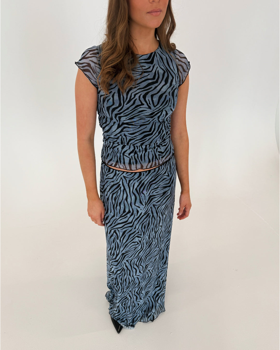 En person iført en lang, blå kjole med zebra-print står i et lyst rum. Personen har løsthængende, bølget hår og sorte sko.