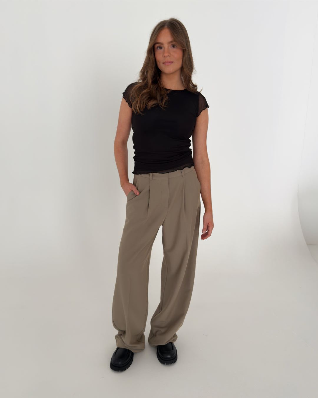 Kvinden står afslappet iført en sort t-shirt og beige bukser. Hun har én hånd i lommen og befinder sig i et minimalistisk, hvidt rum.