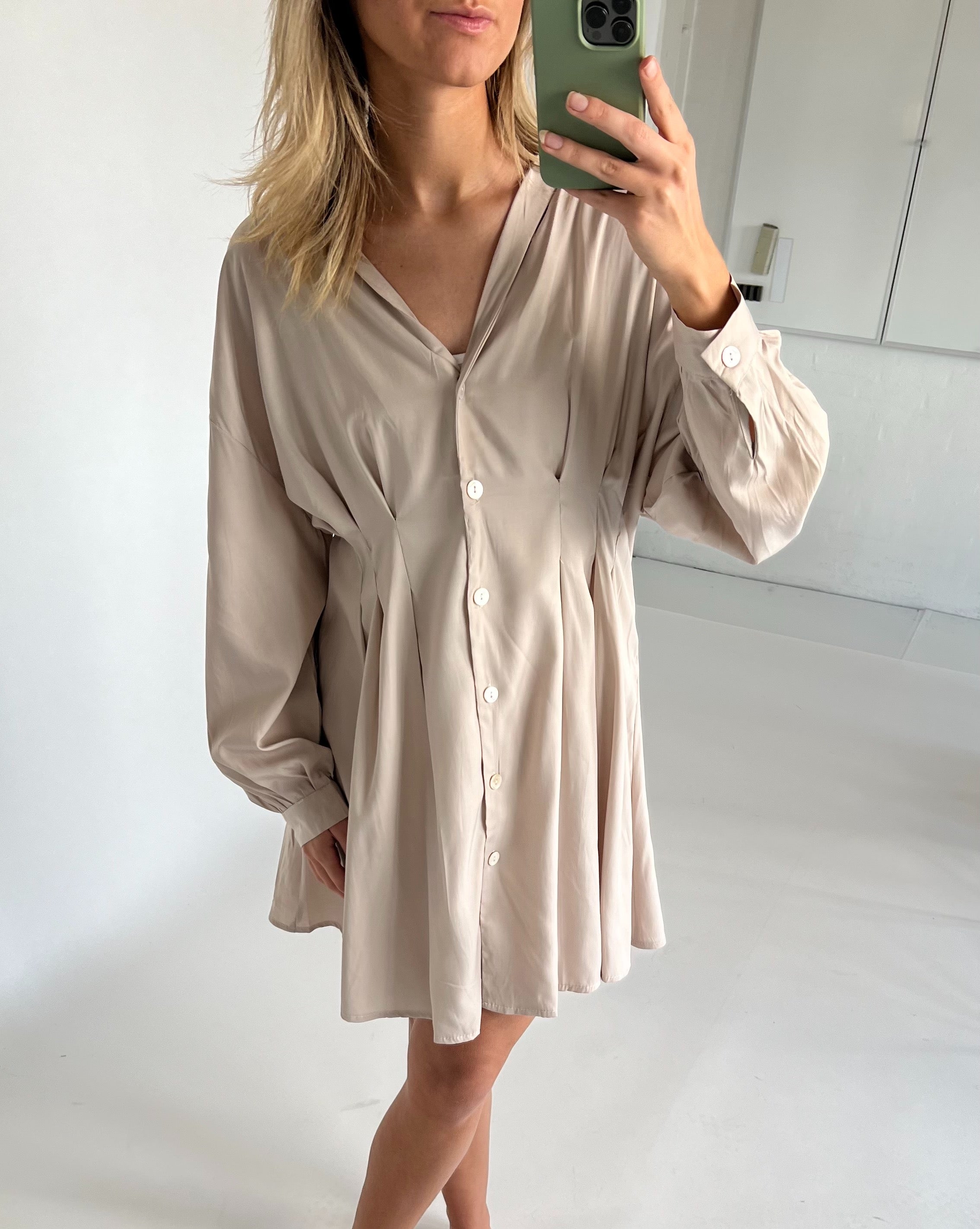 En person iført en beige kjole tager en selfie med en smartphone i et lyst rum. Kjolen har knapper foran og brede ærmer.