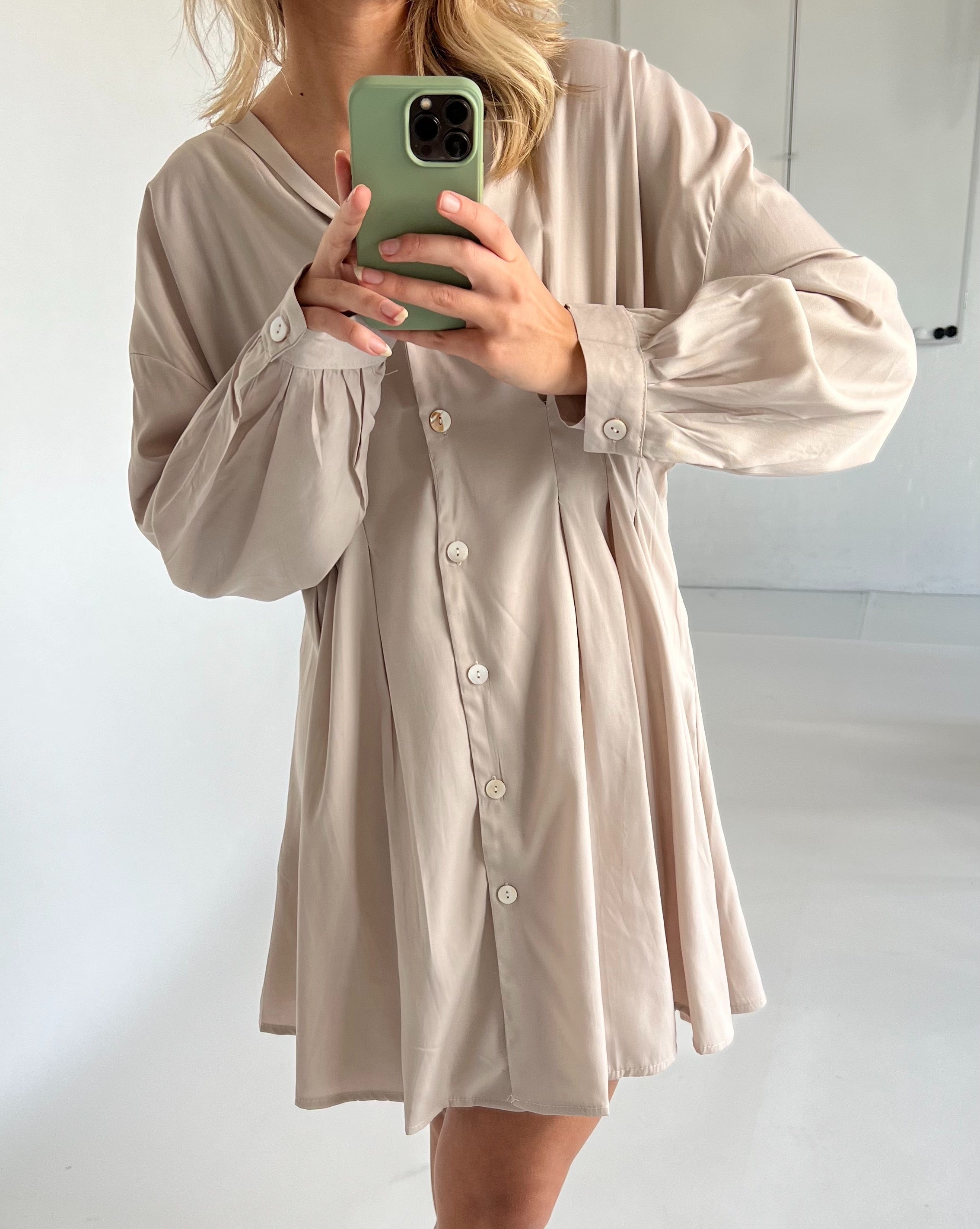 En person iført en beige kjole med knapper fotograferer sig selv i et rum med hvide vægge. Personen holder en mobiltelefon med et grønt cover.