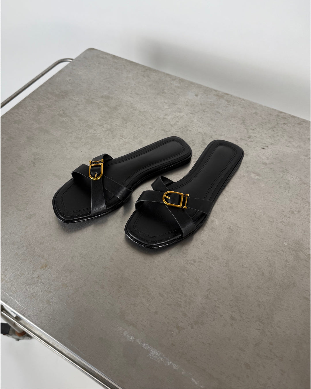 Et par sorte sandaler med guldfarvede spænder ligger stille på en metallisk overflade i et minimalistisk rum.