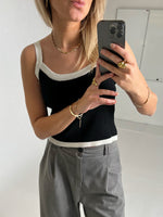 En person tager en selfie i et spejl, iført en sort top og grå bukser, iført guldsmykker og holder en sort smartphone, i et lyst rum uden tekst.
