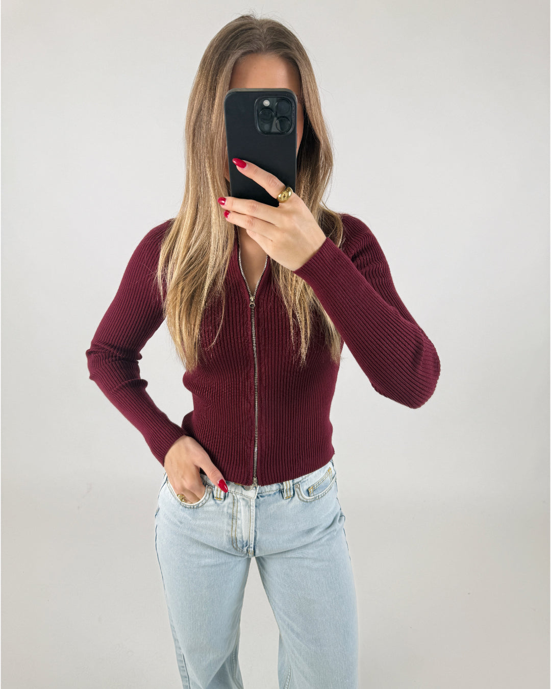 En person iført en bordeaux trøje tager et spejlfoto med en smartphone. Personen har lyse jeans, og i baggrunden er der en ensfarvet, lys væg.