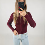 En person iført en bordeaux trøje tager et spejlfoto med en smartphone. Personen har lyse jeans, og i baggrunden er der en ensfarvet, lys væg.