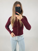 En person iført en bordeaux trøje tager et spejlfoto med en smartphone. Personen har lyse jeans, og i baggrunden er der en ensfarvet, lys væg.