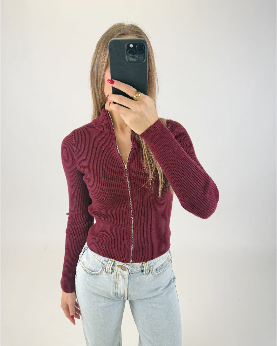 En person iført en bordeaux sweater med lynlås og lyse jeans tager et spejlbillede med en sort telefon i et enkelt, hvidt rum.