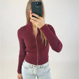 En person iført en bordeaux sweater med lynlås og lyse jeans tager et spejlbillede med en sort telefon i et enkelt, hvidt rum.