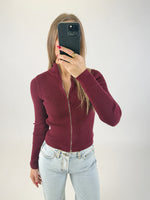 En person iført en bordeaux sweater med lynlås og lyse jeans tager et spejlbillede med en sort telefon i et enkelt, hvidt rum.