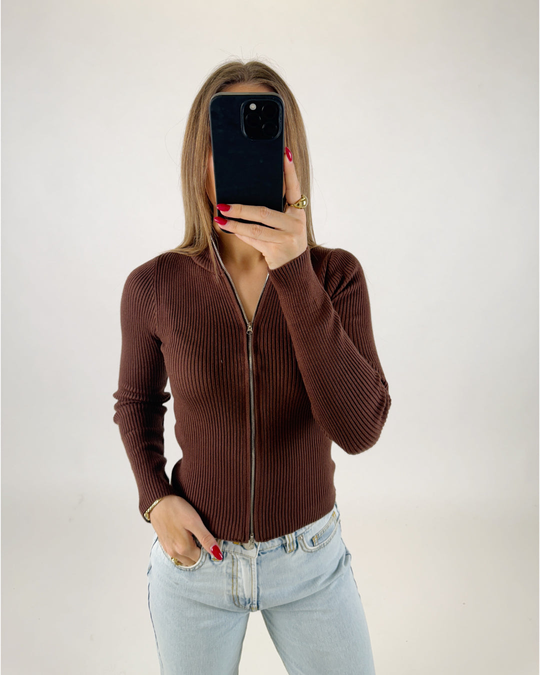 Person holder en mobiltelefon foran ansigtet, iført en brun strikket cardigan og lyseblå jeans, i et neutralt belyset rum. Røde negle og guldringe er synlige.