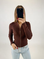 Person holder en mobiltelefon foran ansigtet, iført en brun strikket cardigan og lyseblå jeans, i et neutralt belyset rum. Røde negle og guldringe er synlige.