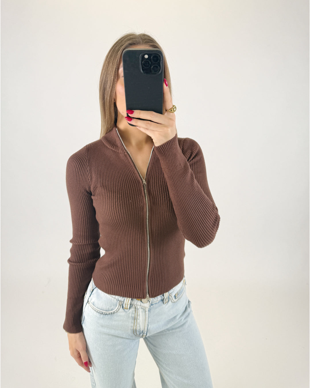 En person tager et selfie iført en brun trøje med lynlås i en lys, minimalistisk baggrund, der dækker ansigtet med en sort mobiltelefon.
