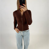 En person står foran et spejl, tager en selfie med en telefon. Personen bærer en brun lynlåsbluse og lyseblå jeans, omgivet af en neutral baggrund.