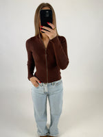 En person står foran et spejl, tager en selfie med en telefon. Personen bærer en brun lynlåsbluse og lyseblå jeans, omgivet af en neutral baggrund.