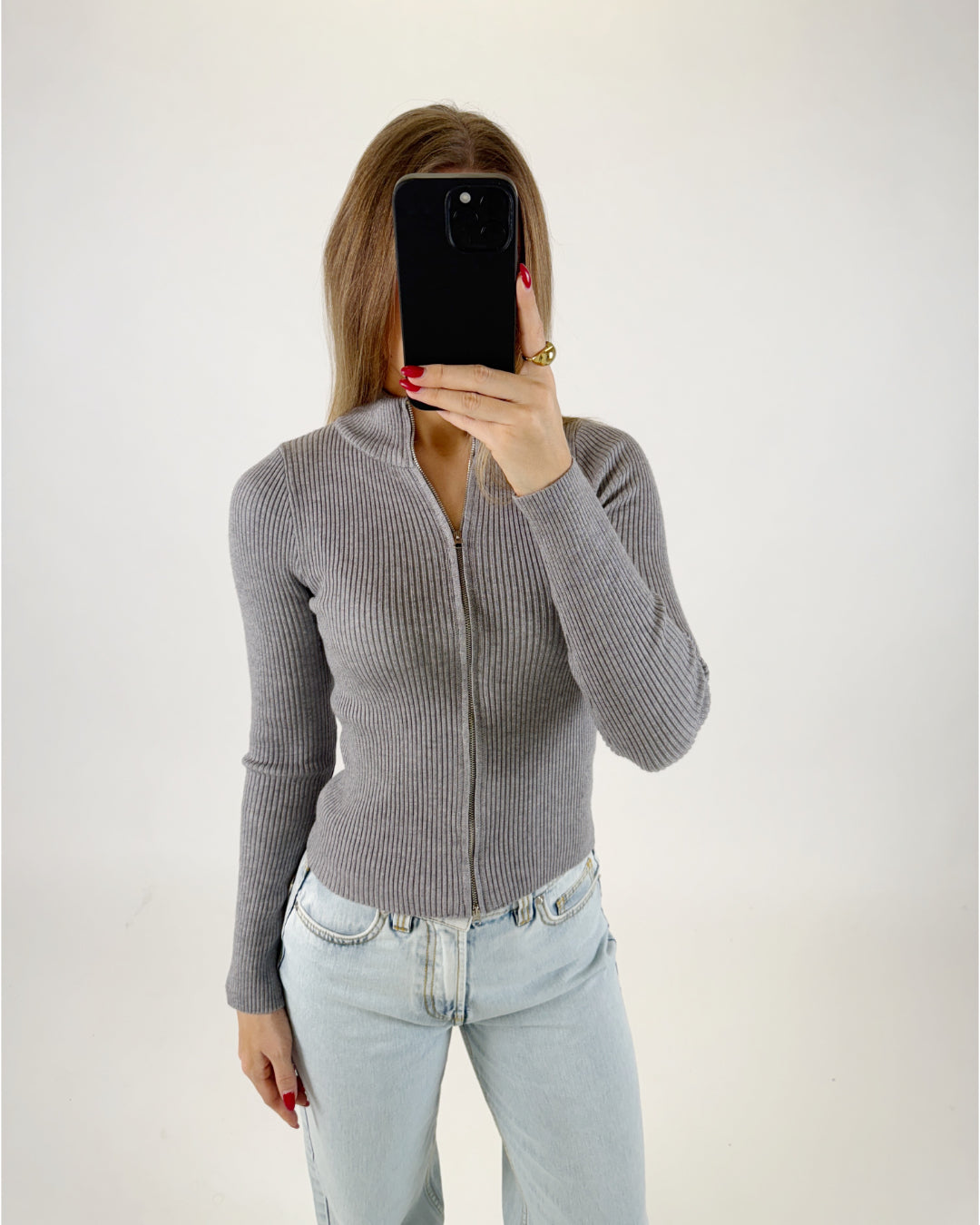 En person iført en grå, tætsiddende trøje og lyse jeans holder en sort mobiltelefon foran ansigtet, i et neutralt, lysegråt rum.
