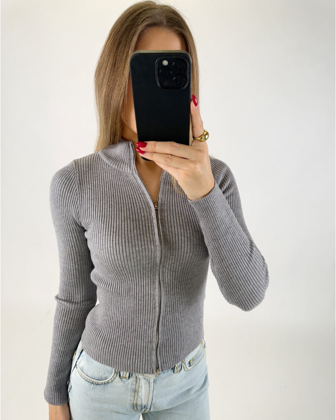En person holder en mobiltelefon foran ansigtet, iført en grå striktrøje med lynlås og lyseblå jeans. Baggrunden er hvid og enkel.
