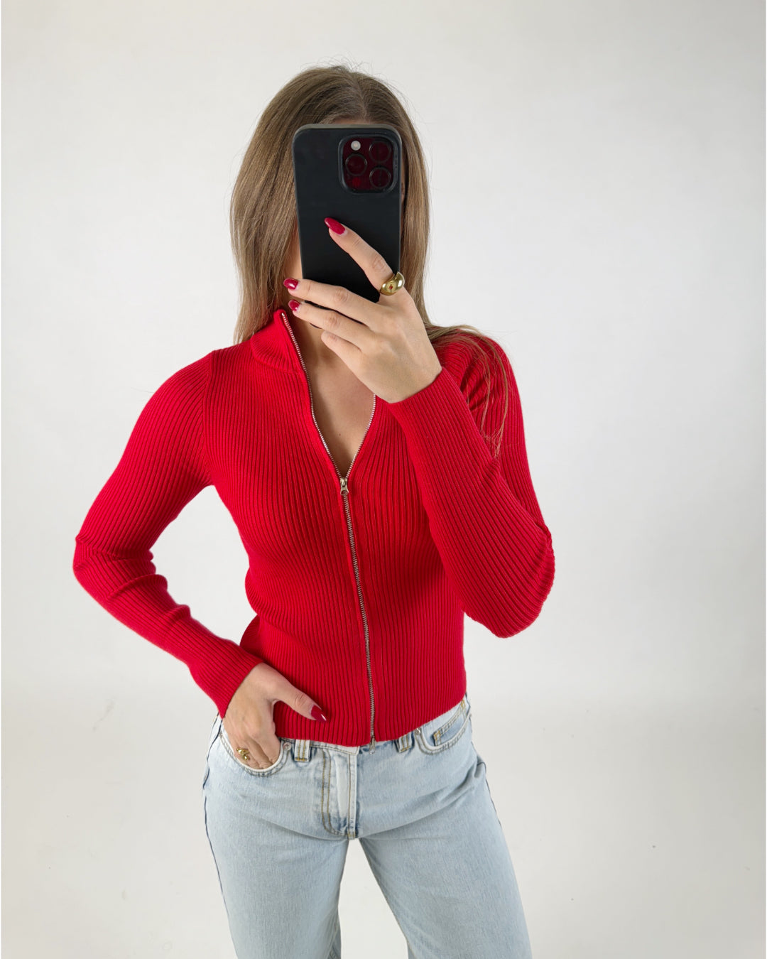 En person i rød trøje tager en selfie med en sort telefon i en lys baggrund. Personen har hånden i en bukselomme og bærer smykker.