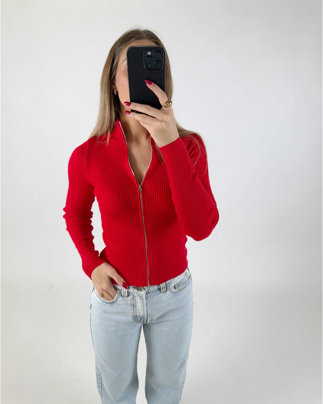 En person i en rød lynlås-trøje tager et billede af sig selv med en sort mobiltelefon. Personen står i et lyst, neutralt rum og bærer blå jeans.