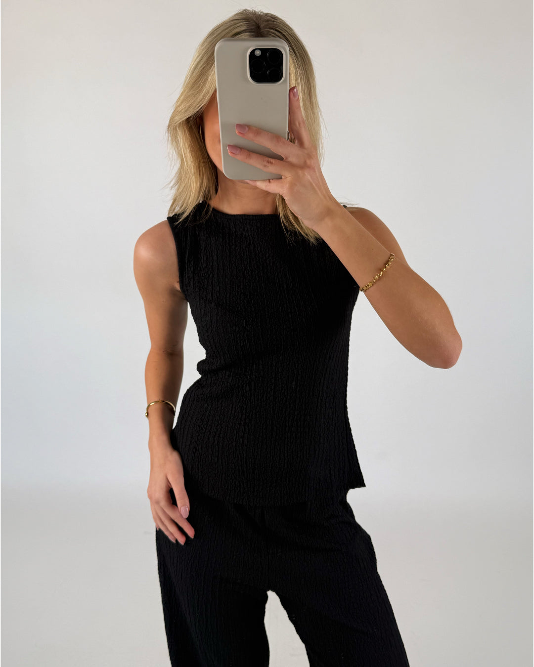 En person iført sort ærmeløs top tager en selfie foran et blankt væg. Hånden holder en mobiltelefon, og der er et armbånd på håndleddet.