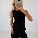En person iført sort ærmeløs top tager en selfie foran et blankt væg. Hånden holder en mobiltelefon, og der er et armbånd på håndleddet.